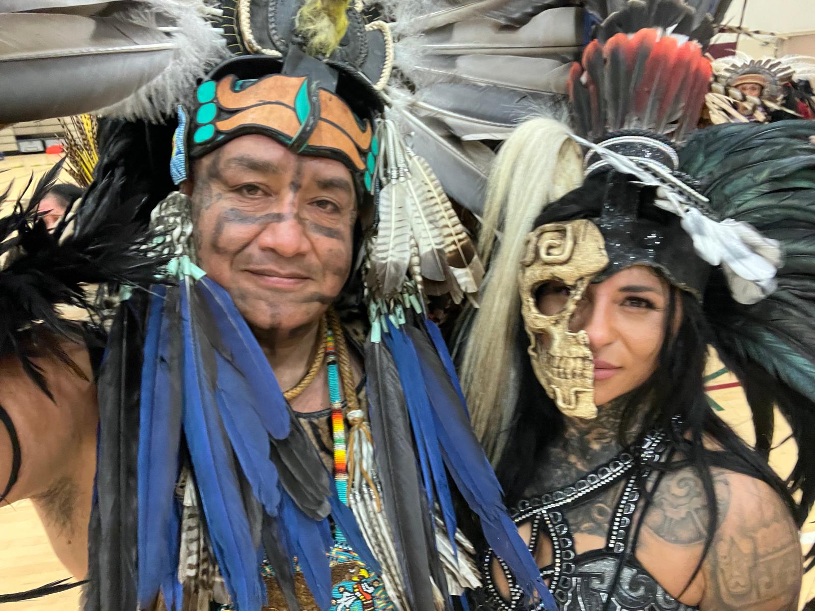 Two Aztec dancers in feathered regalia — one with face paint, one with skull mask — posing together