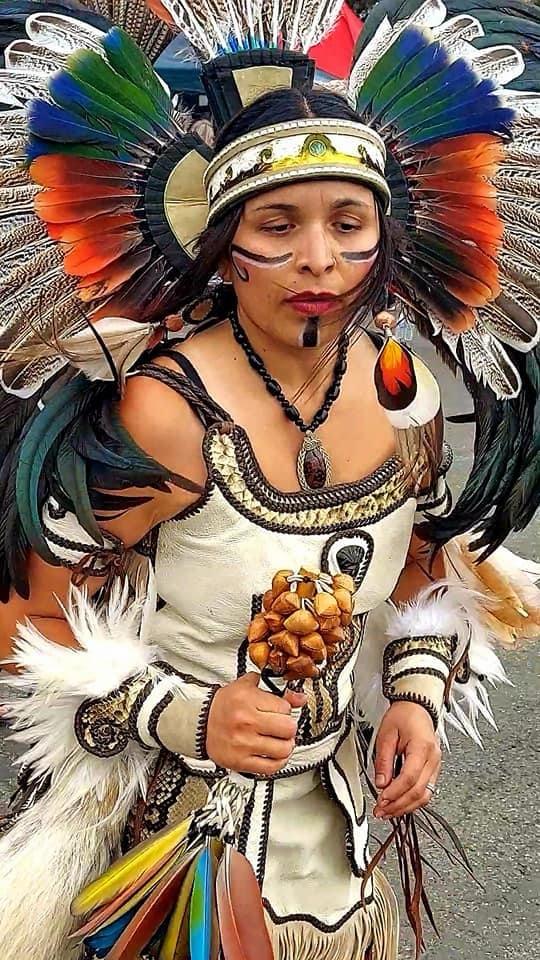 Female Aztec dancer in white regalia with colorful feathered headdress holding a rattle