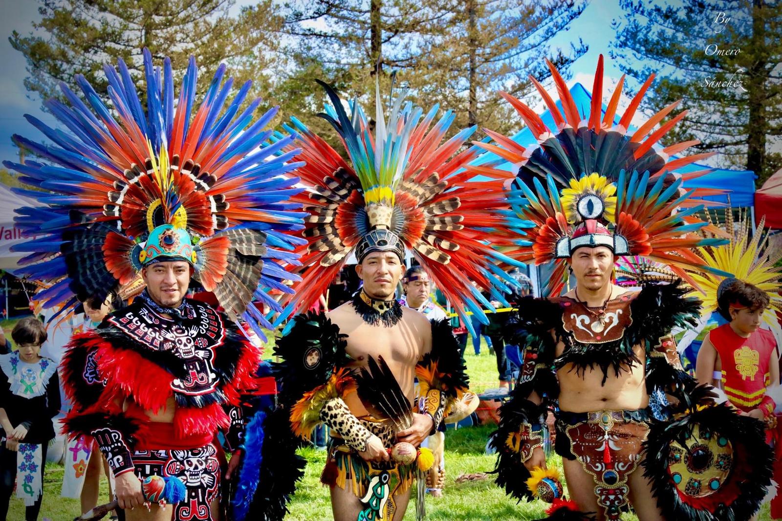 Three Aztec dancers in vibrant multicolored feathered headdresses posing together