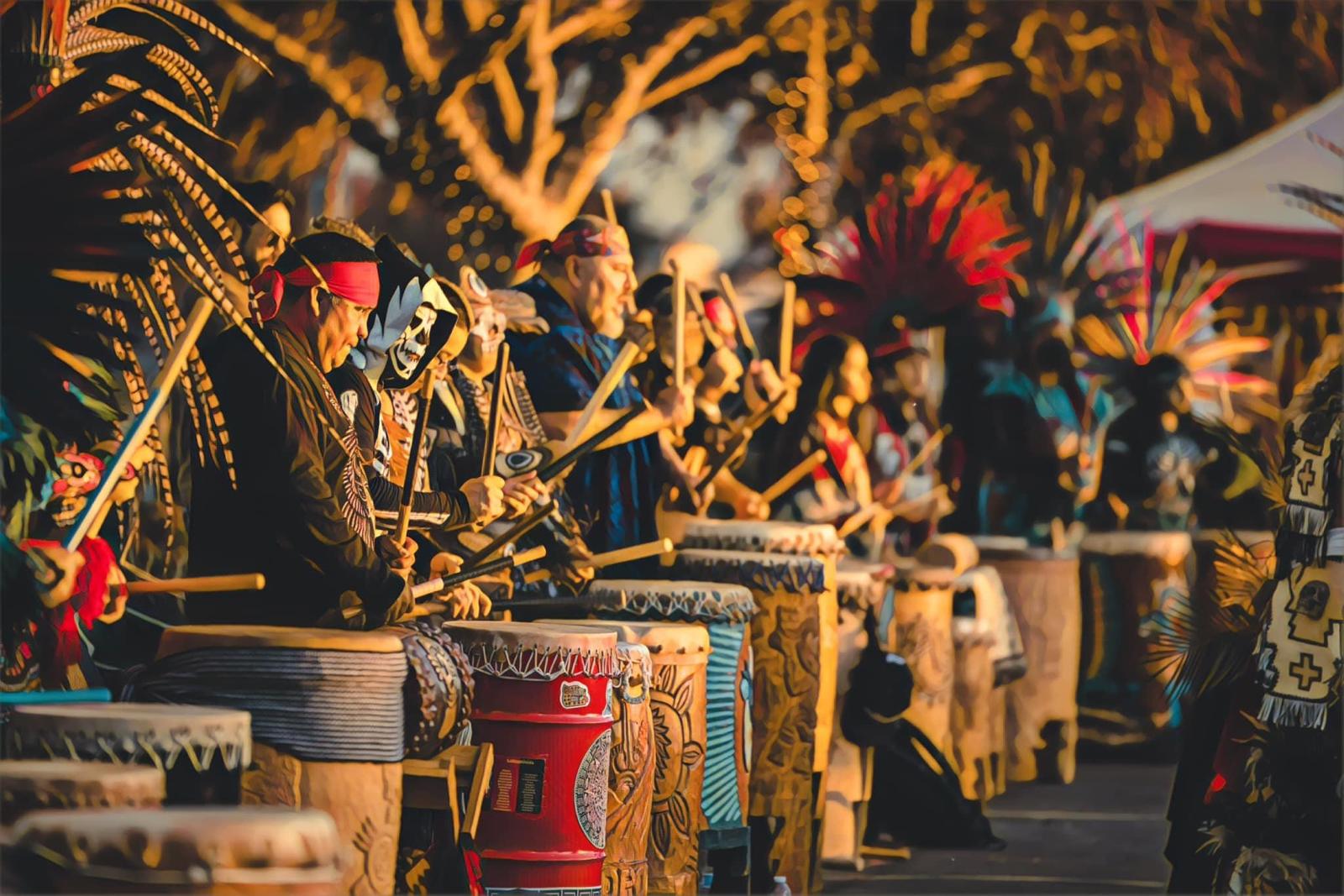 Row of Aztec drummers in feathered regalia and skull masks playing ceremonial drums at golden hour