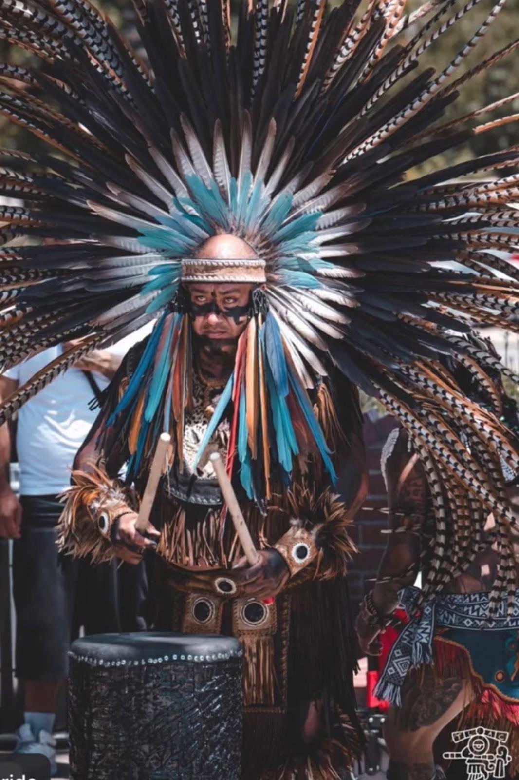 Aztec warrior in large blue and brown feathered headdress playing ceremonial drum