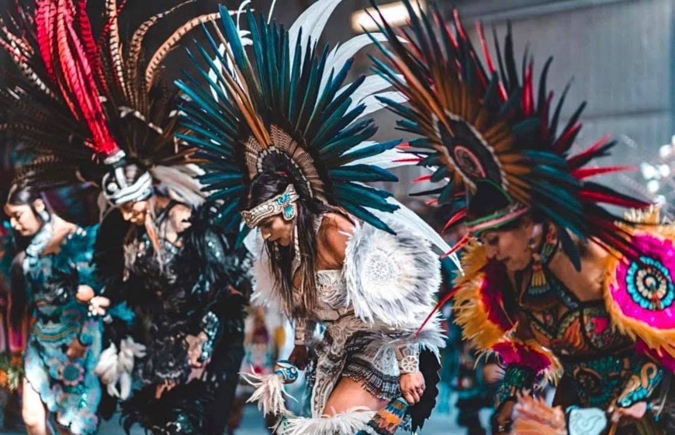 Three female Aztec dancers in teal and colorful feathered headdresses mid-performance