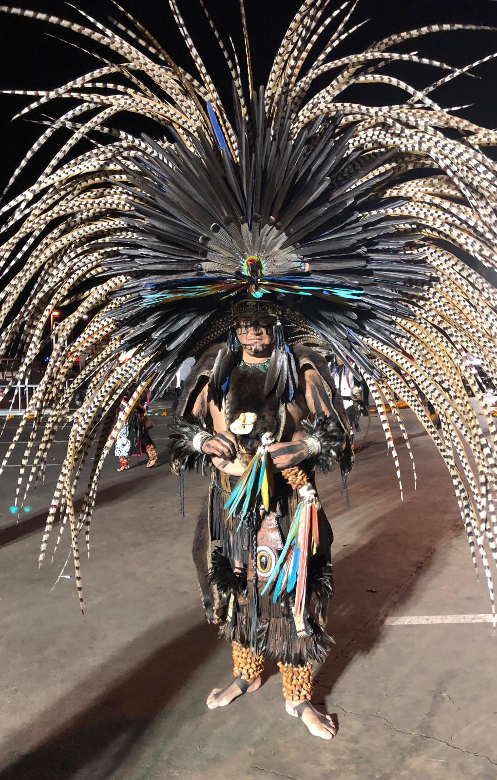 Aztec dancer in large black and teal feathered headdress standing barefoot at night