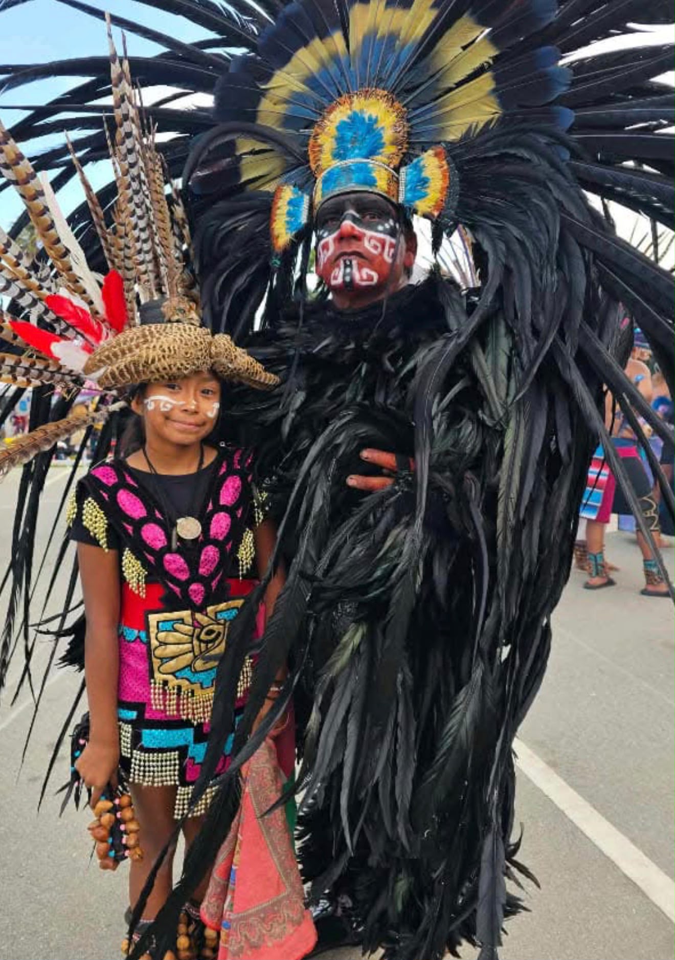 Young girl in colorful Aztec regalia with feathered hat and face paint standing beside a tall dancer in full black feathered headdress and ceremonial face paint